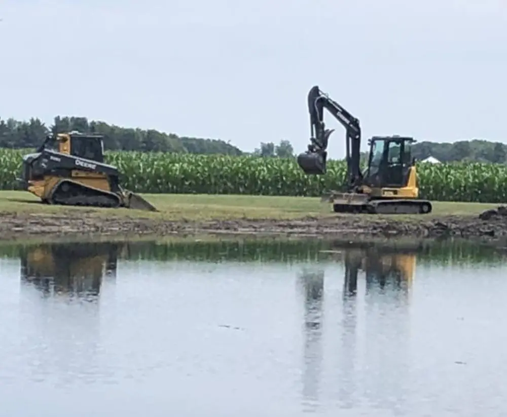 Two machines shaping a pond basin with a cornfield beyond